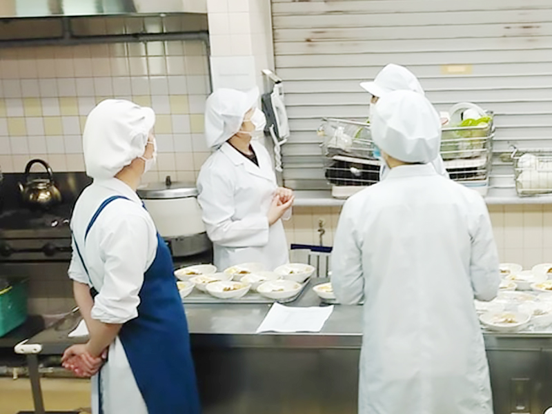 セントラルキッチンの現場でスタッフが集まり盛り付けの準備や作業確認を行っている様子